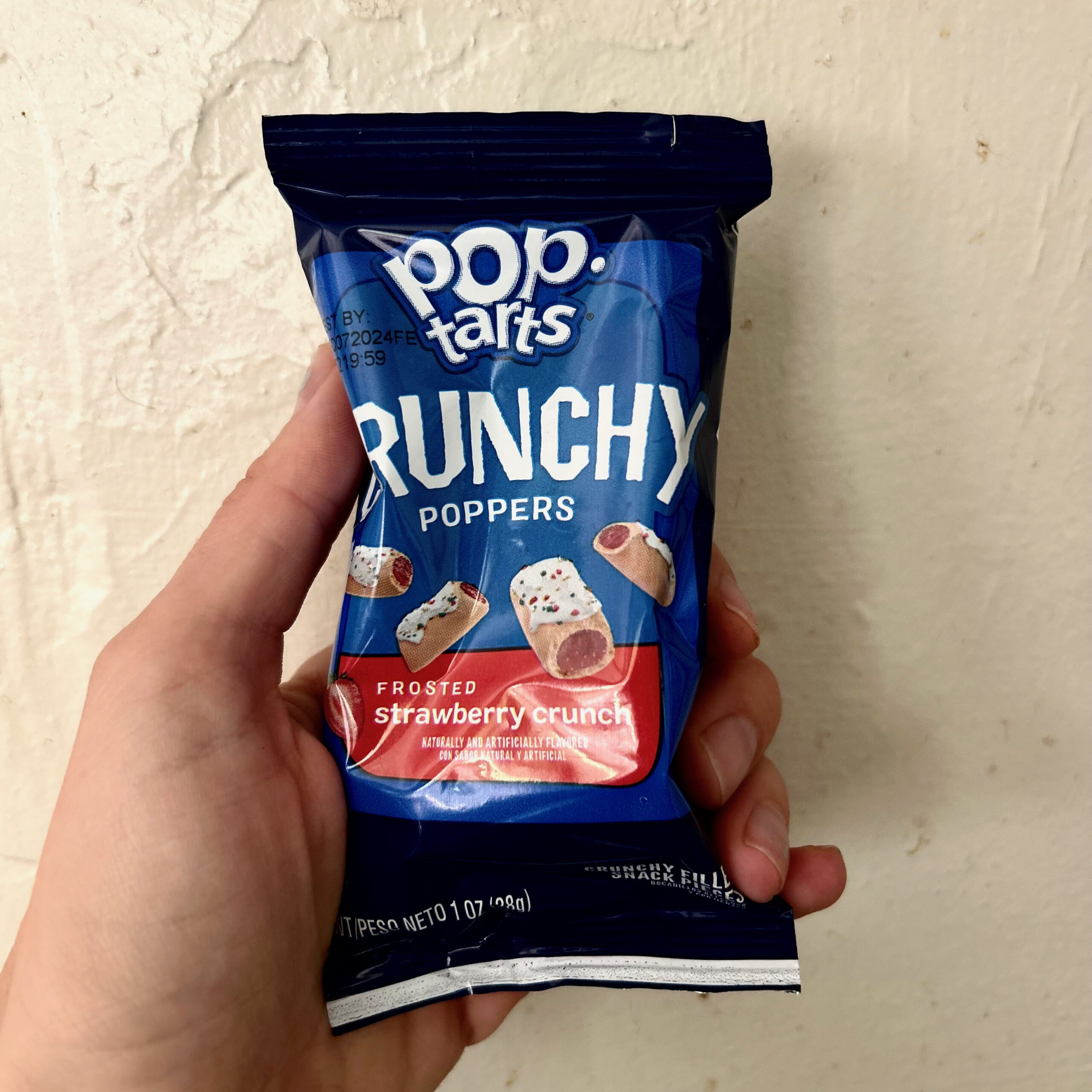 A hand holding a packet of Pop-Tarts Crunchy Poppers. The package is blue and shows pink pod-shaped foods filled with red goo.